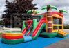 A colorful inflatable bounce house with slides and palm tree decorations, set up outdoors on a blue ground cover in a parking lot under a cloudy sky.