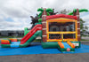 A tropical-themed inflatable bounce house and slide set up outdoors on a blue ground cover. The bounce house features green palm trees, brown faux-trunk pillars, and a red and yellow windowed bouncy area, with a green and red slide on the left and orange steps in front.