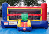 A large inflatable bounce house with a small slide, photographed outdoors on a blue tarp with trees and a fence in the background. The bounce house is primarily blue, red, and yellow, with a green slide section.