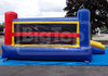 A large inflatable bouncy castle with a blue base and walls, red side pillars, a yellow-trimmed front play panel, and a small yellow slide on the right, photographed outdoors on a blue tarp over asphalt with trees and sky in the background.