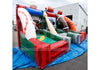 An inflatable baseball-themed bounce house with a slide, featuring red, green, white, and orange sections, set up outdoors on a blue tarp in front of a white building.