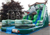 A large inflatable dinosaur gator slide with a green and blue jungle-themed design, featuring a gray dinosaur climbing down the slide. The inflatable is set up outdoors on a paved surface with trees in the background.