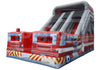 An inflatable fire truck themed obstacle course or bounce house with a slide, featuring red, white, and blue graphics, yellow hazard stripes, and gray inflatable walls.
