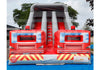 An inflatable fire truck themed slide bounce house with two red fire truck fronts, a gray slide, and yellow-and-red chevron accents, photographed outdoors on a blue ground cover.