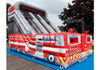 An inflatable fire truck themed bouncy castle with a large slide, set outdoors on a blue floor mat with trees and a cloudy sky in the background.