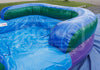 An inflatable swimming pool with a blue water-filled interior, green inflatable walls, and purple side supports, photographed outdoors on a blue tarp with grass visible in the background.