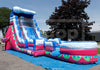 A large inflatable water slide playset with a pink and blue design, set up outdoors on a blue ground cover with trees and a building in the background.