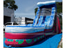 An inflatable water slide playground with a blue and white slide, a pink and red body, and green accents, set outdoors on a blue tarp with buildings and trees in the background.