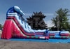 A large inflatable water slide bounce house with a pink, blue, and white design, set up outdoors on a blue tarp over asphalt with trees and a blue sky in the background.