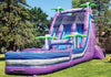 A large inflatable water slide playground with purple, blue, and white sections, green palm tree graphics, and a black top canopy, set up on a grassy lawn with trees in the background.